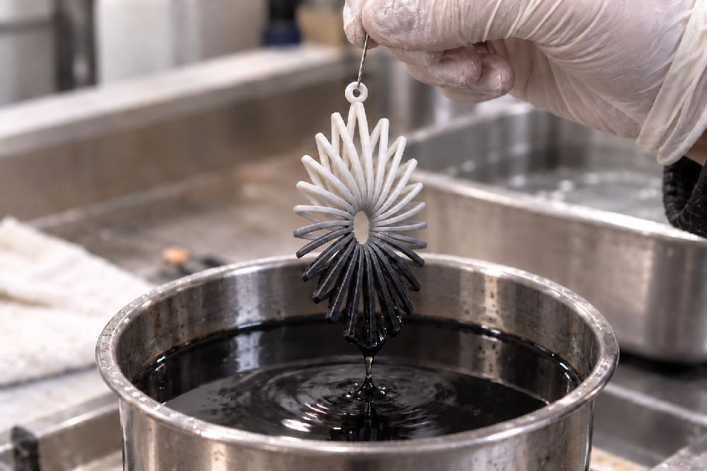 A white 3D printed nylon pendant lifted from a bath of black dye, dyed below the waterline and still bone white above it
